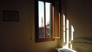 Un dormitorio con cama, mesita de noche y ventana con vista a un edificio con torre.