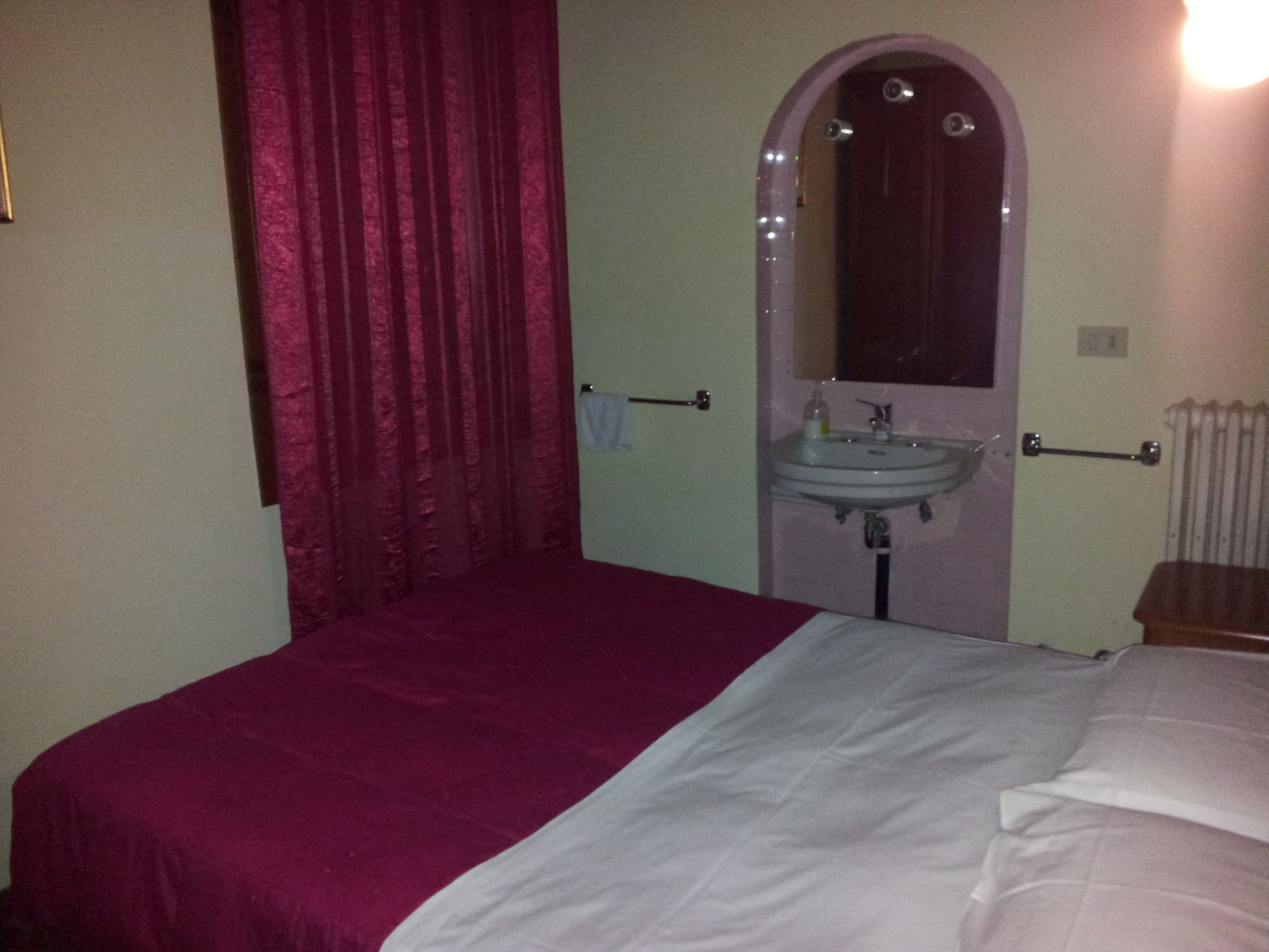 A bedroom with a bed, a red curtain, a white bedspread, a white wall, a white door, a white radiator, and a white sink with a mirror.