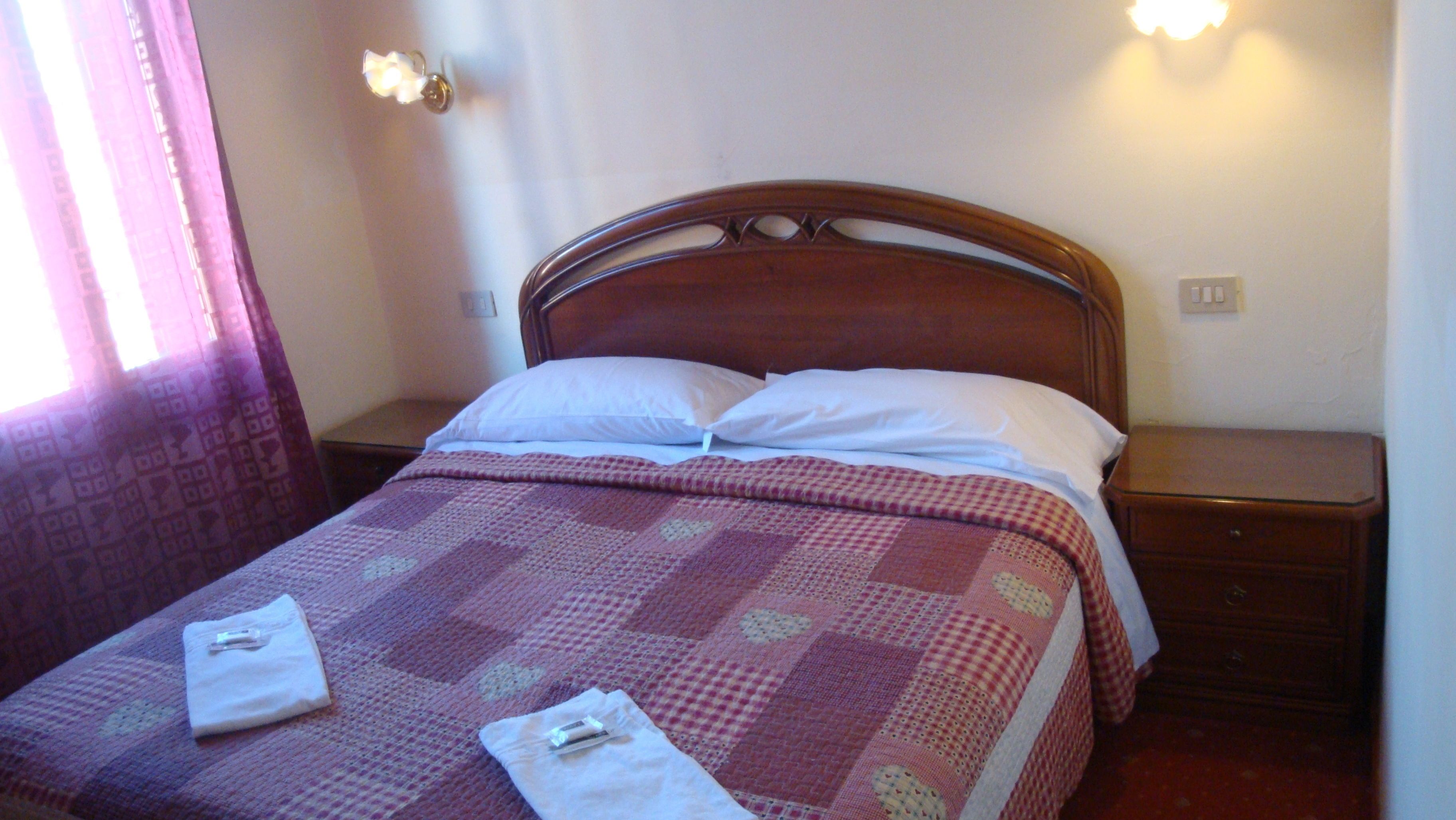 A neatly made bed with a checkered bedspread and two pillows, a wooden headboard, a nightstand with a lamp, and a window with curtains.