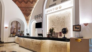 Een hotel receptie met een modern design, met een receptiebalie met een digitaal scherm, een muur met Egyptische hiërogliefen, en een klok.
