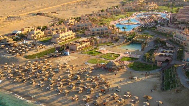 Luchtbeeld van een resort met meerdere gebouwen, een groot zwembadgedeelte en tal van strandparasols.