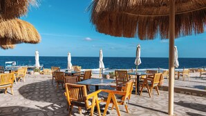 Een café aan het strand met houten tafels en stoelen, rieten parasols en uitzicht op de oceaan.