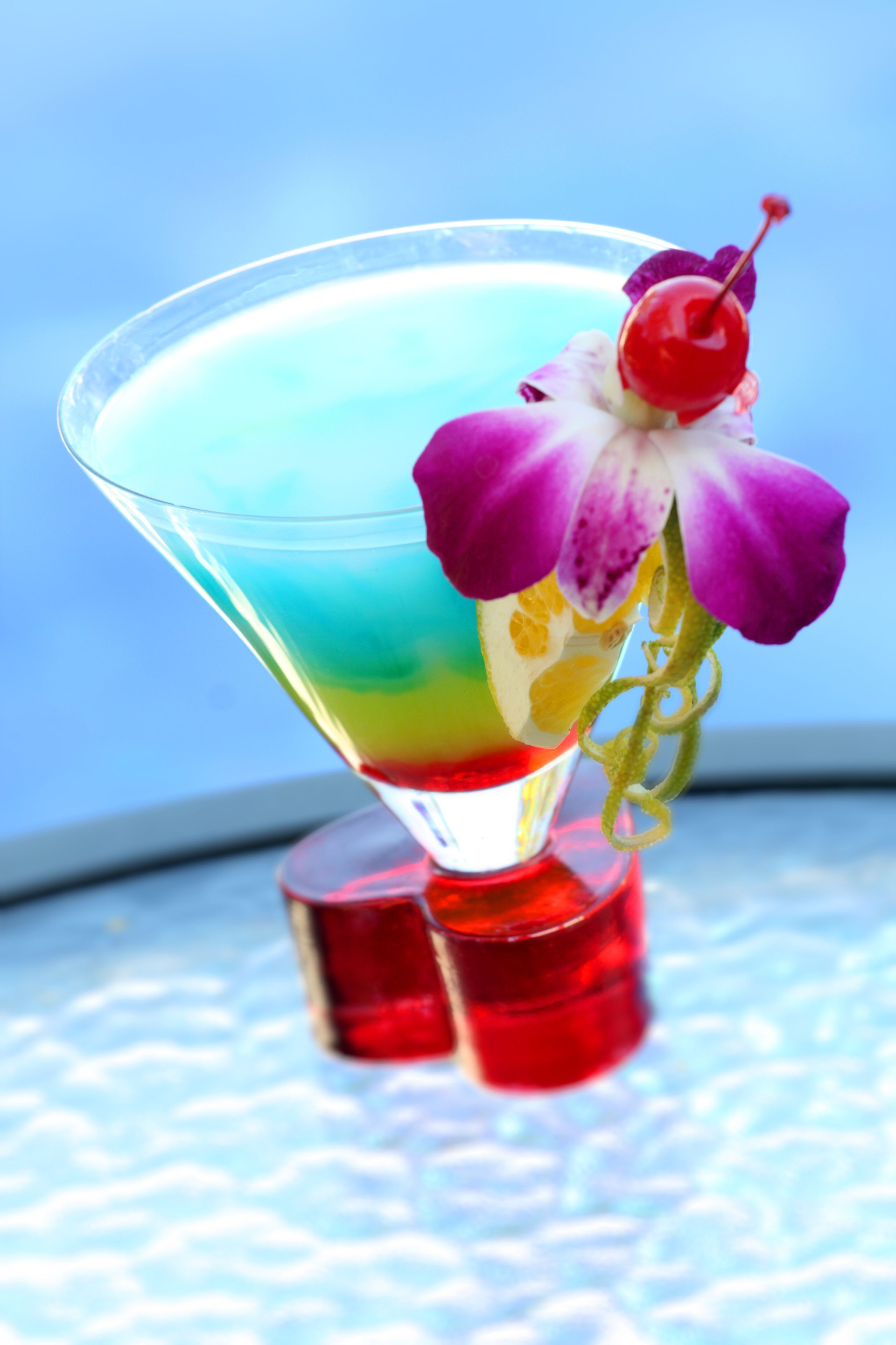 Ein Martini-Glas mit einem schichtartigen Getränk und einer violetten Orchideenblüte auf einer spiegelnden Oberfläche.