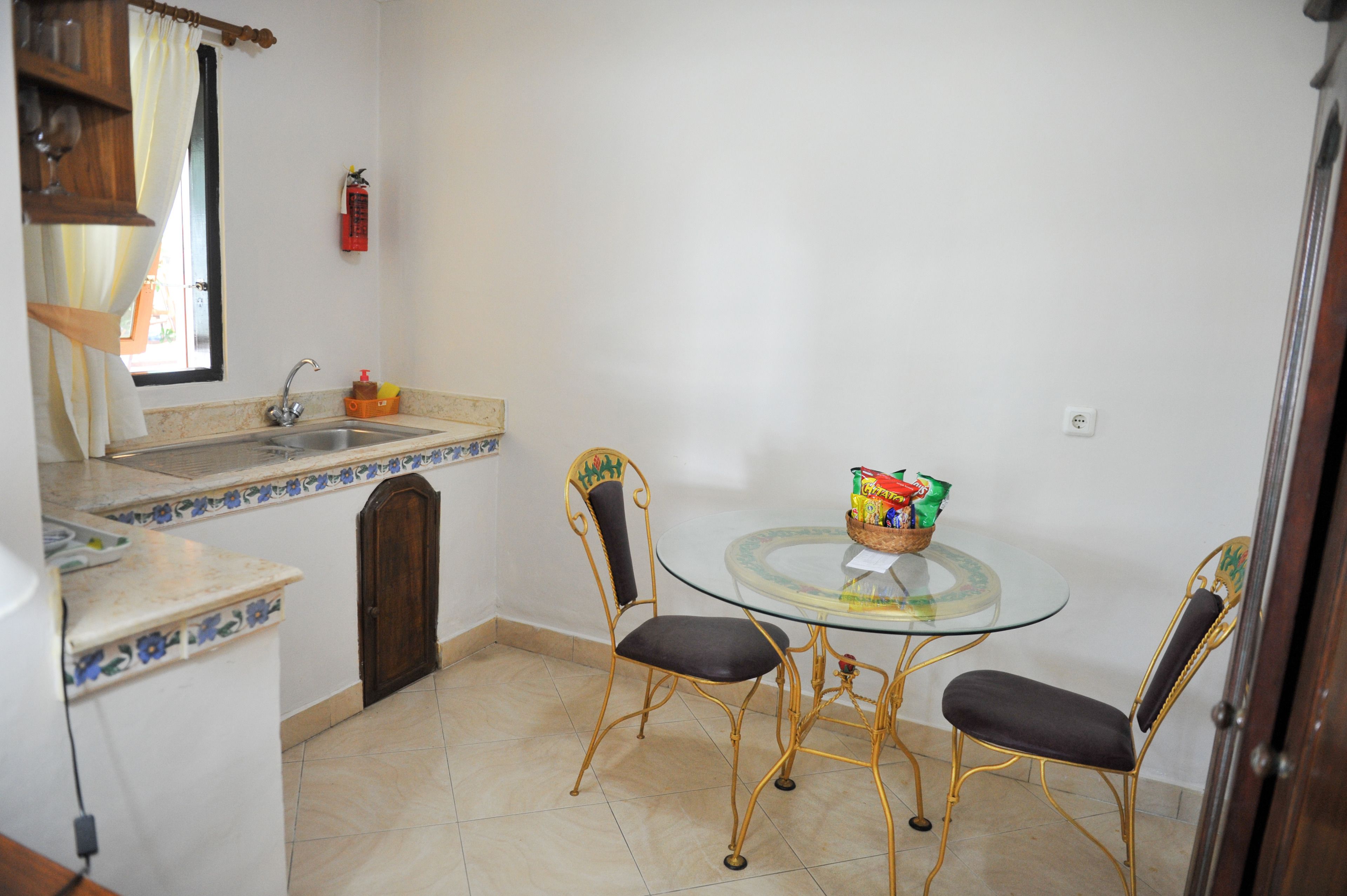 Una pequeña cocina con un lavabo, una ventana con cortinas y una mesa redonda de cristal con dos sillas.