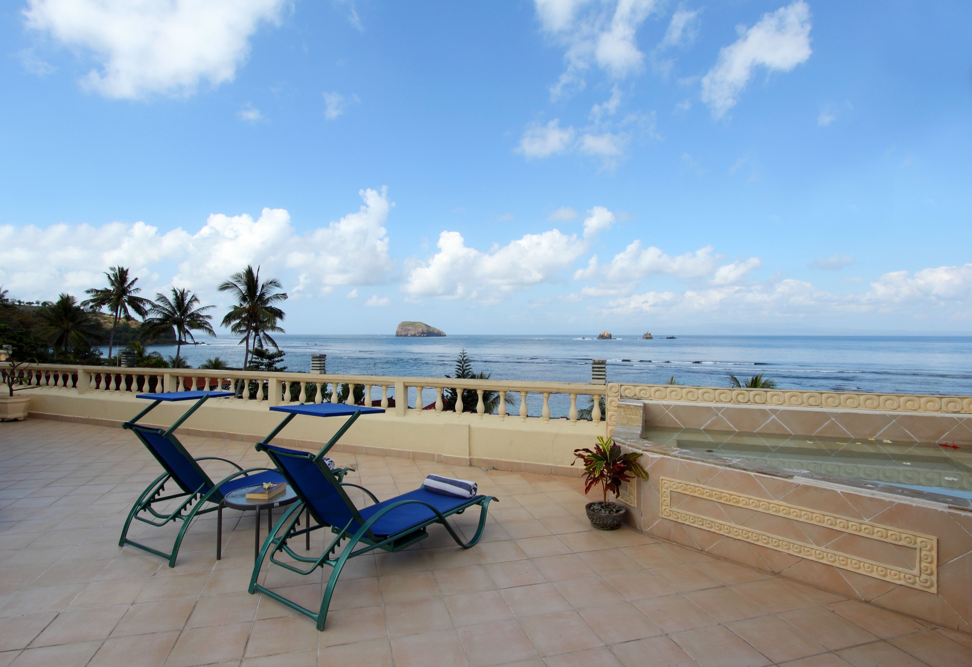 A terrace with two blue lounge chairs and a small table, overlooking a beach with palm trees and the ocean.