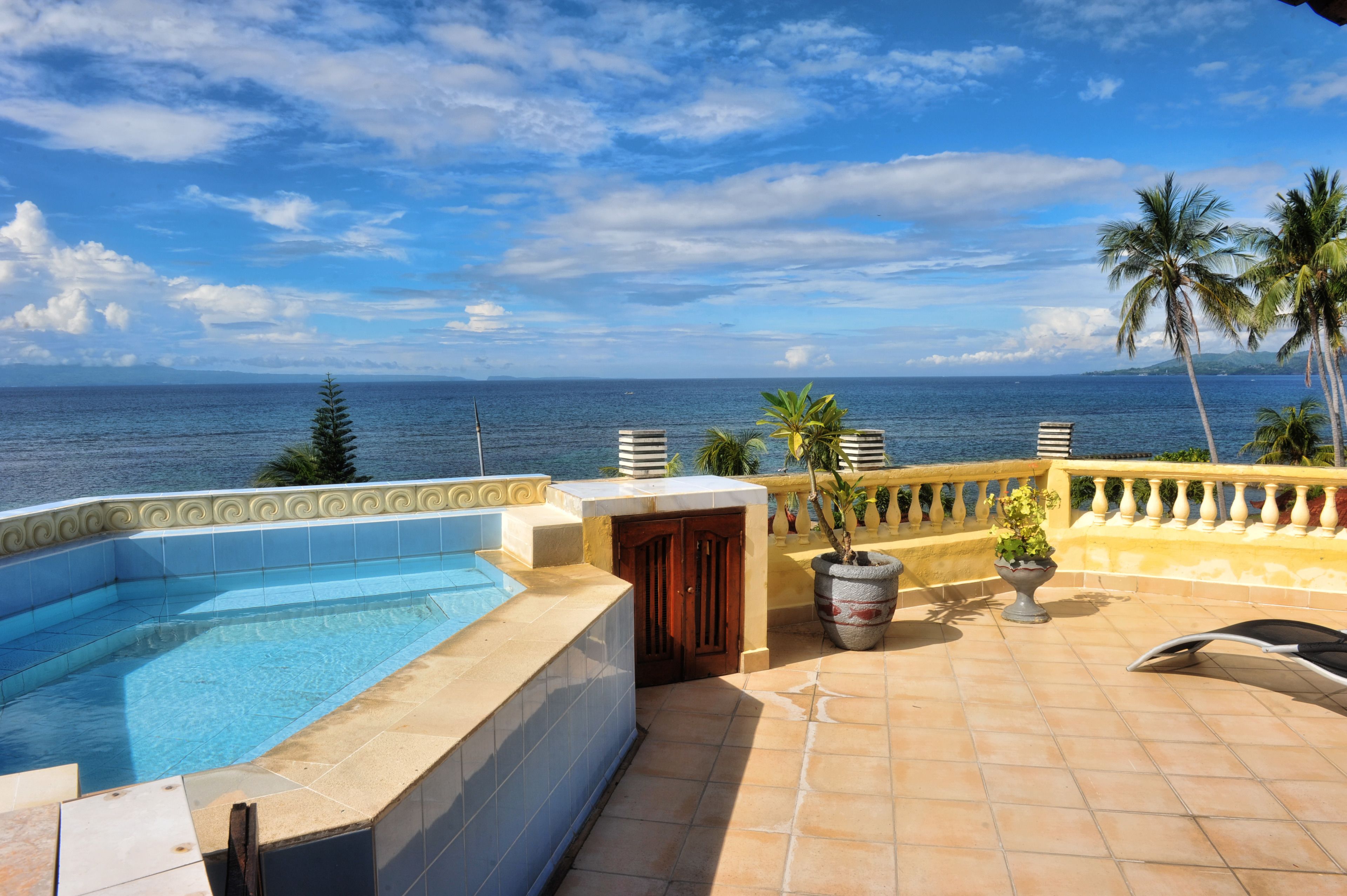 A rooftop pool with a view of the ocean, palm trees, and a tiled deck.