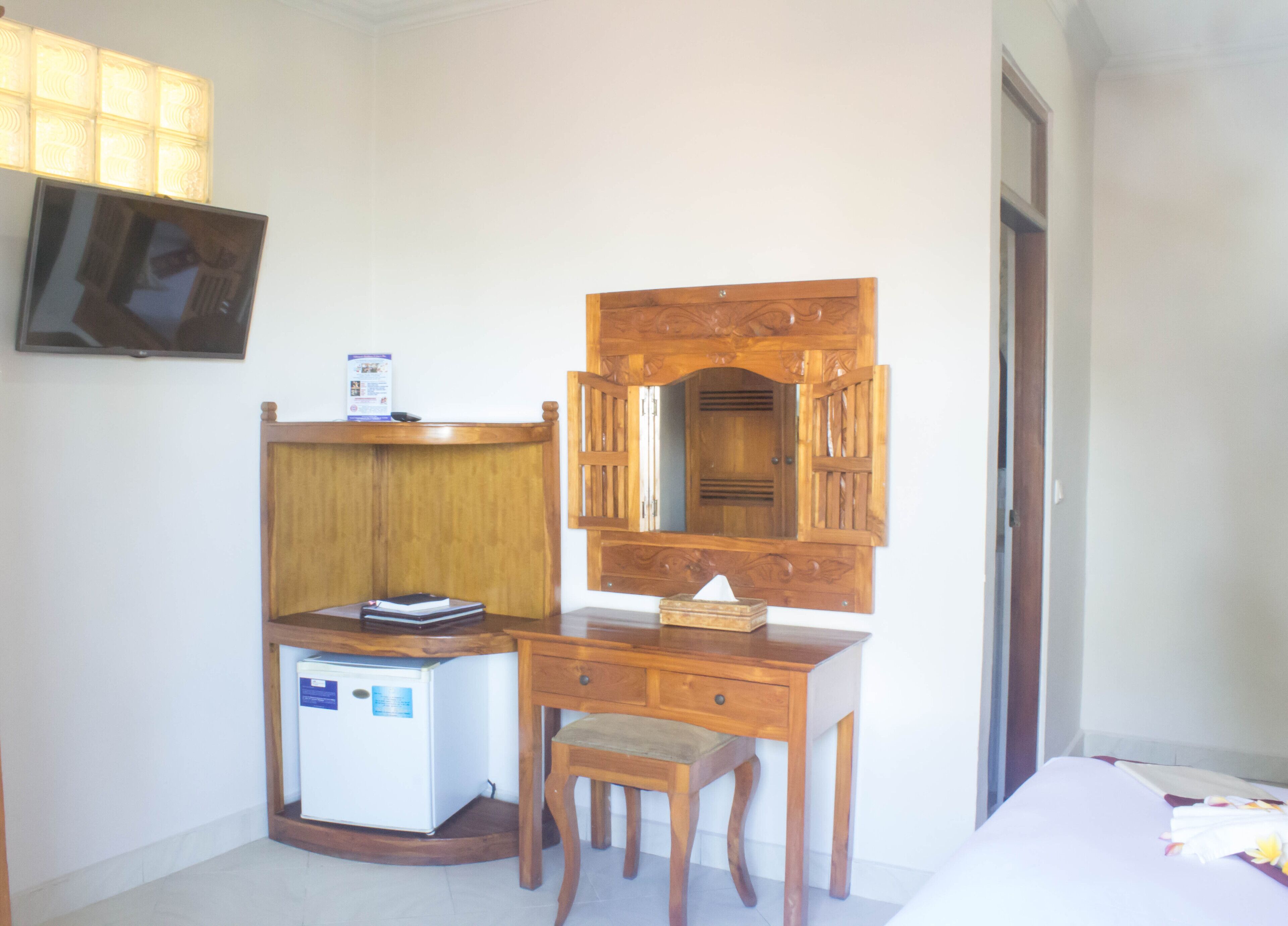 Een kleine, netjes ingerichte slaapkamer met een houten bureau, een stoel, een kleine koelkast, een aan de muur bevestigde televisie en een deur naar een andere kamer.