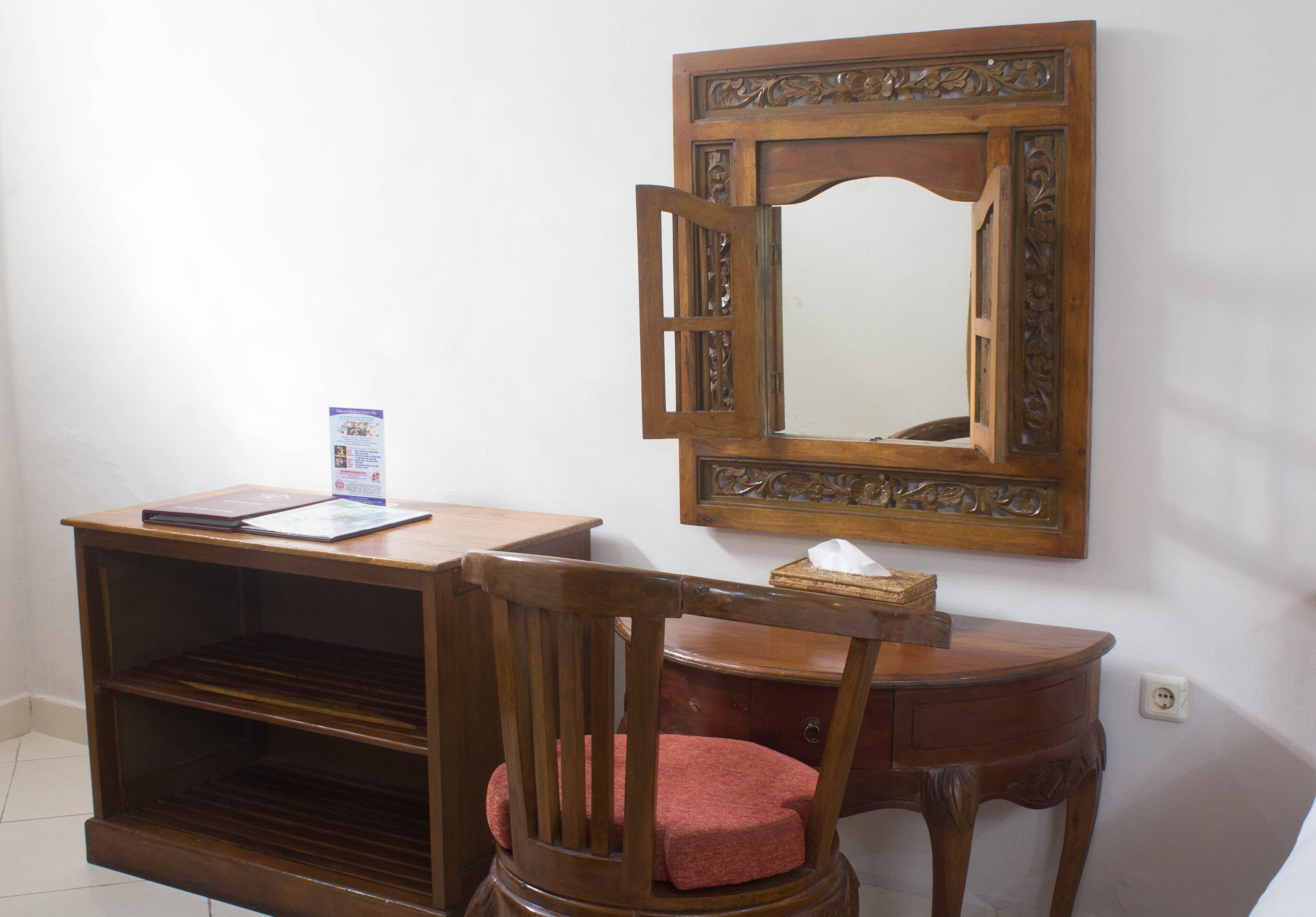 Un escritorio de madera con una computadora portátil y un folleto, una silla con un cojín rojo, y un espejo en la pared.