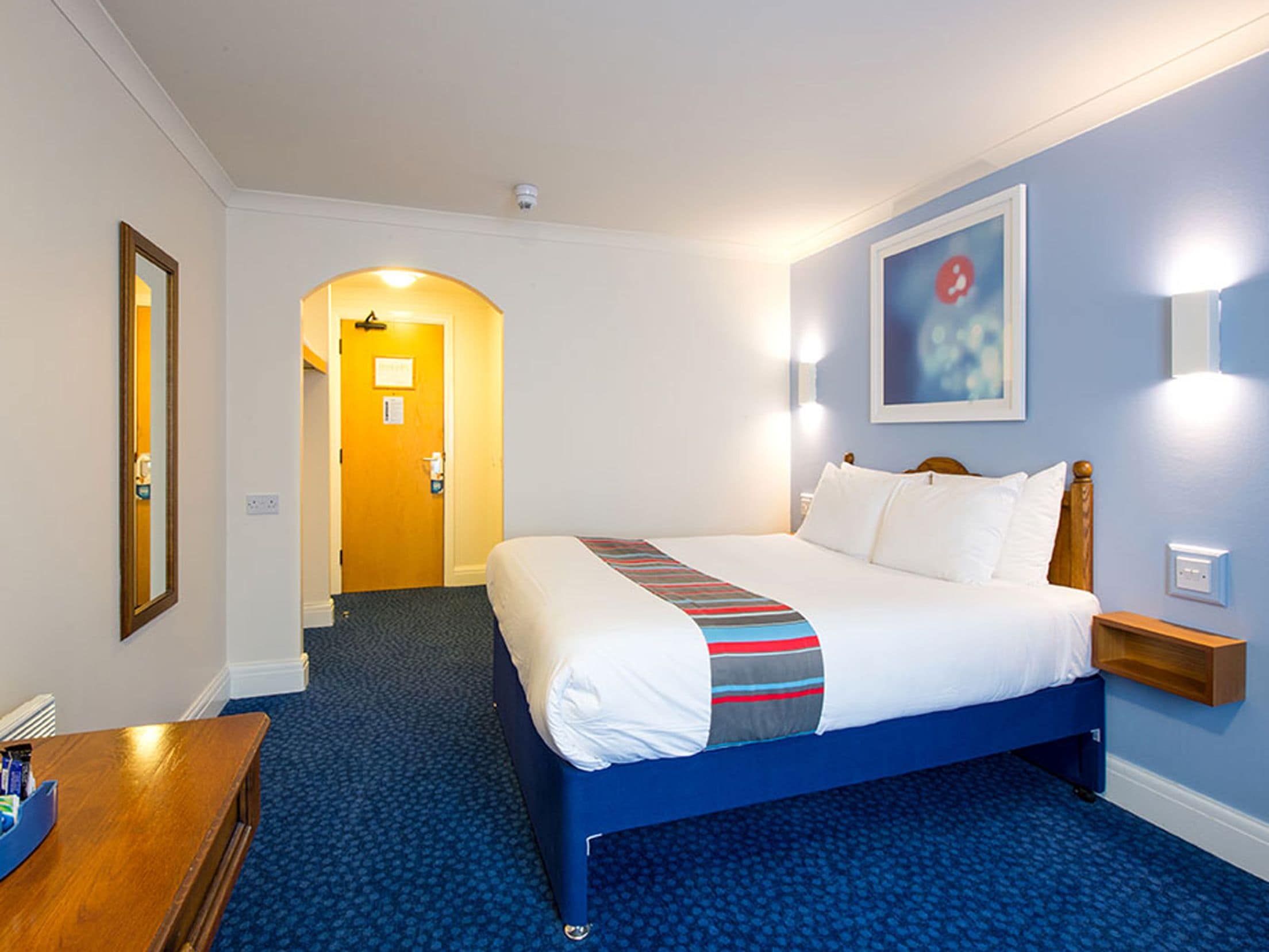 A hotel room with a bed, a desk, a chair, a mirror, a door, and a wall-mounted light fixture.