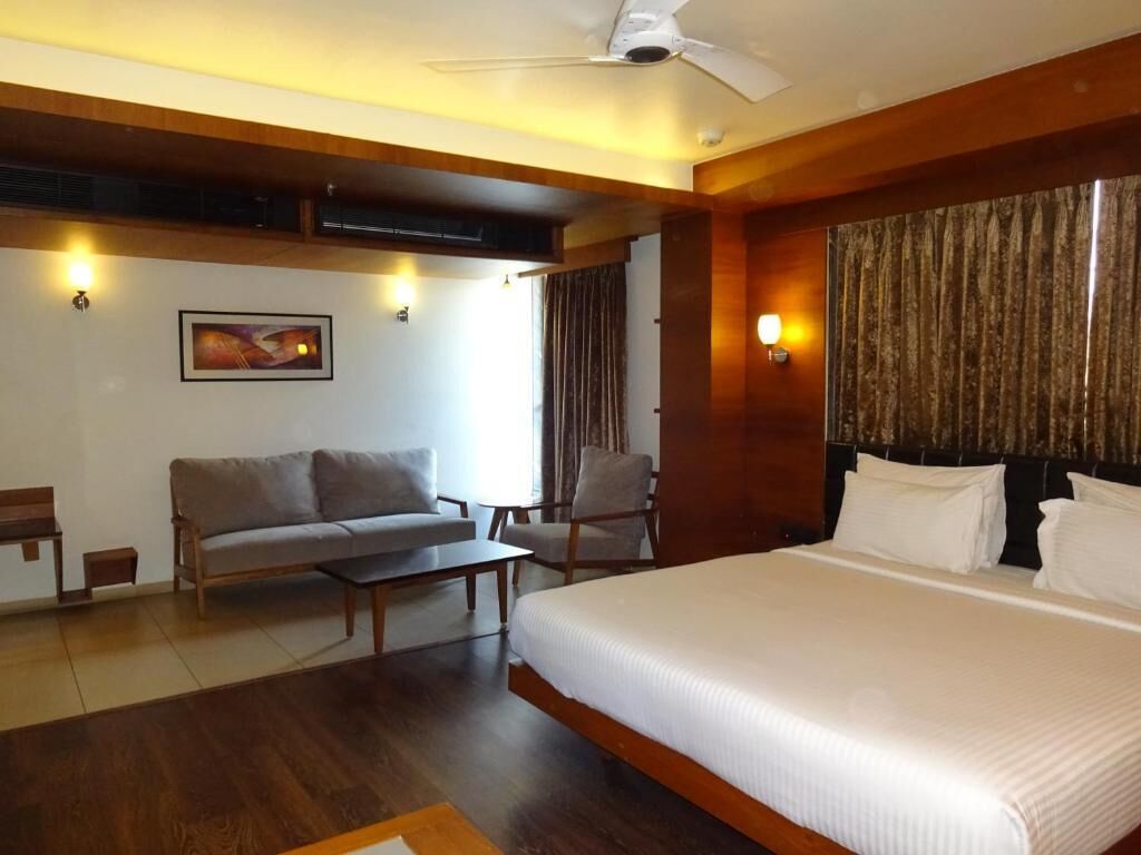 A hotel room with a large bed, a sofa, a chair, and a desk. There is a ceiling fan and a painting on the wall.