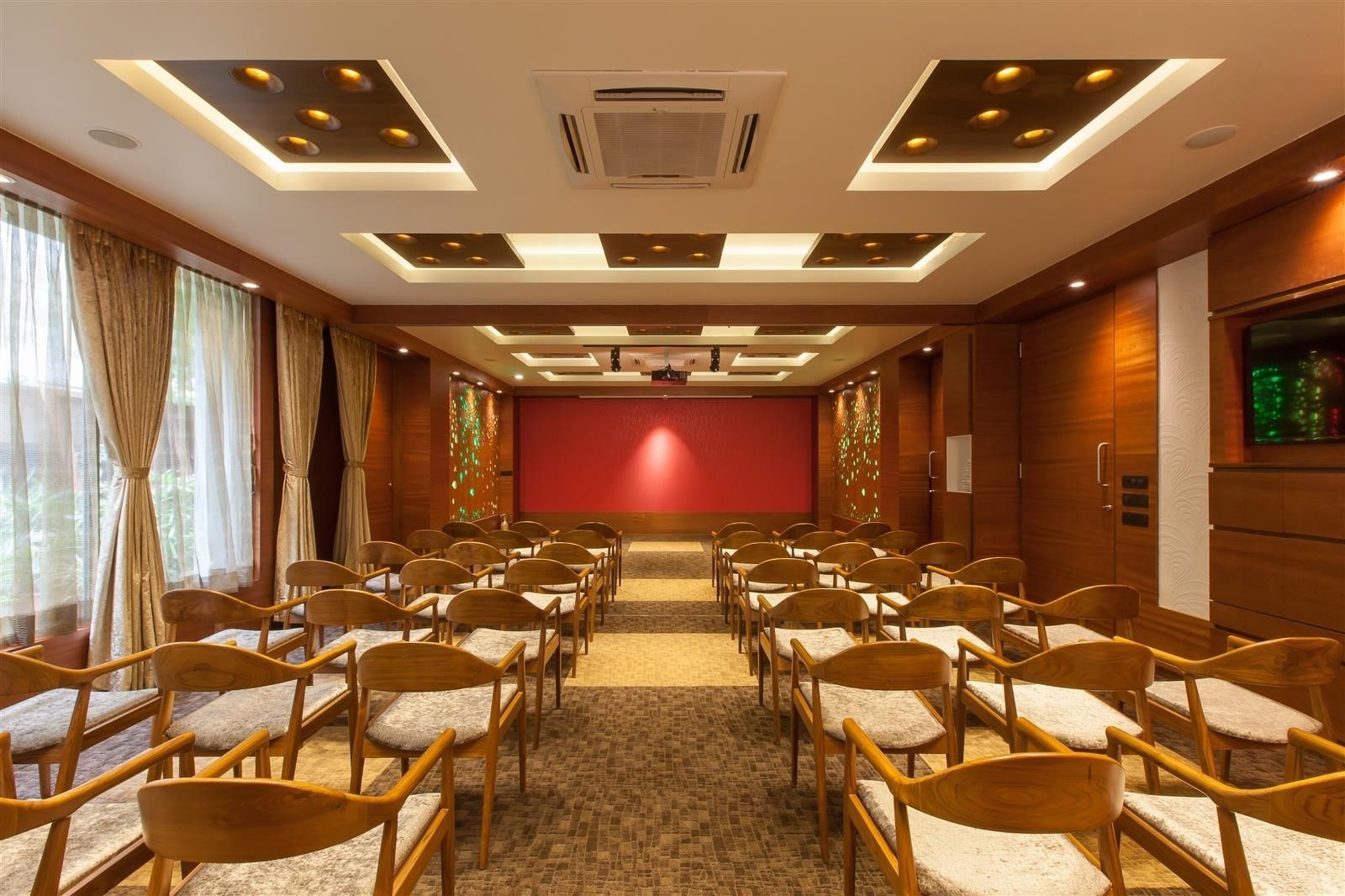 A conference room with rows of wooden chairs, a red screen, and a ceiling with recessed lighting.