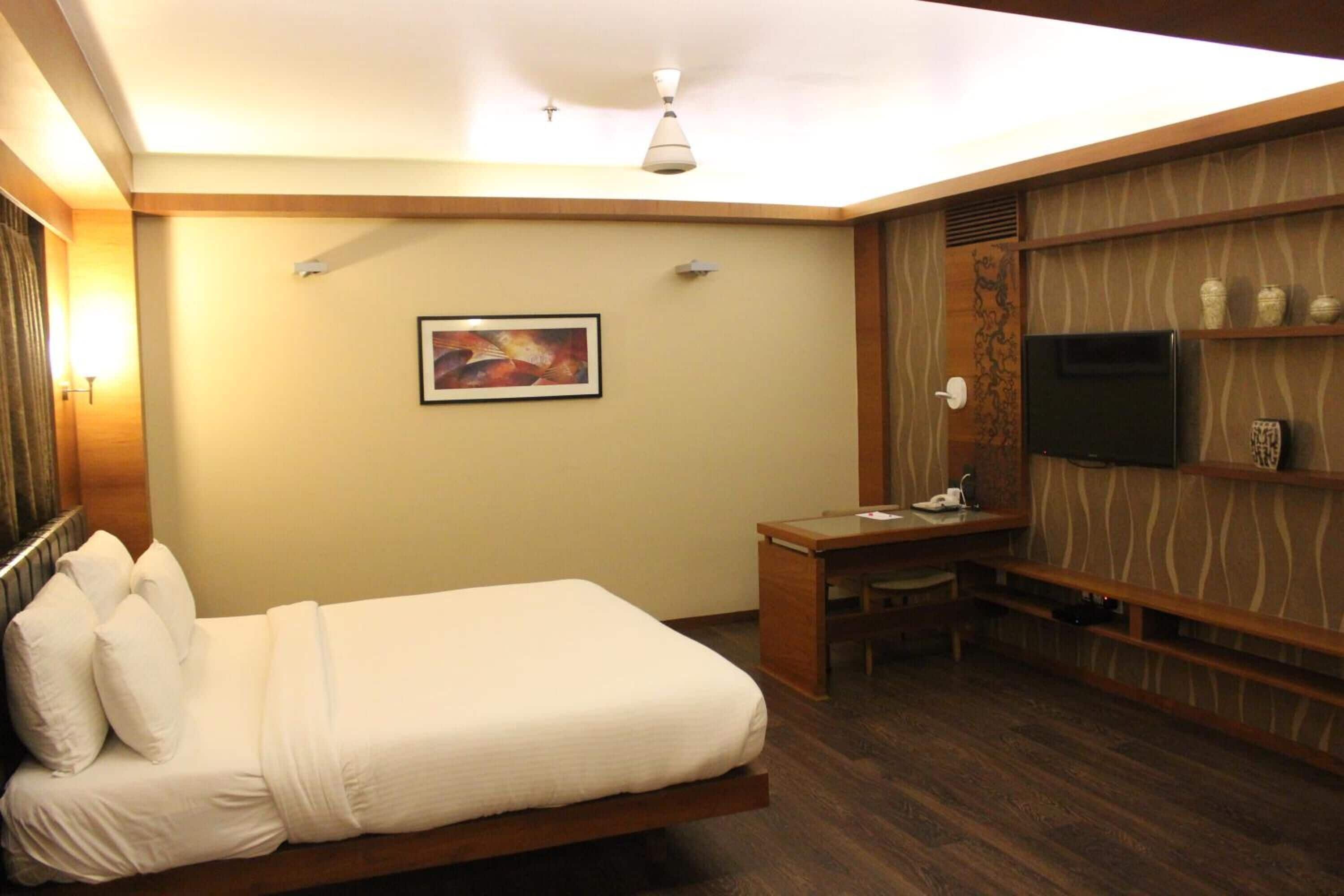 Habitación de hotel con cama, escritorio, silla, televisor y objetos decorativos en estantes.