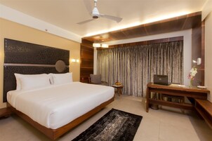 Quarto de hotel com uma cama grande, uma mesa com um laptop, uma cadeira, uma mesinha com frutas e uma cortina.