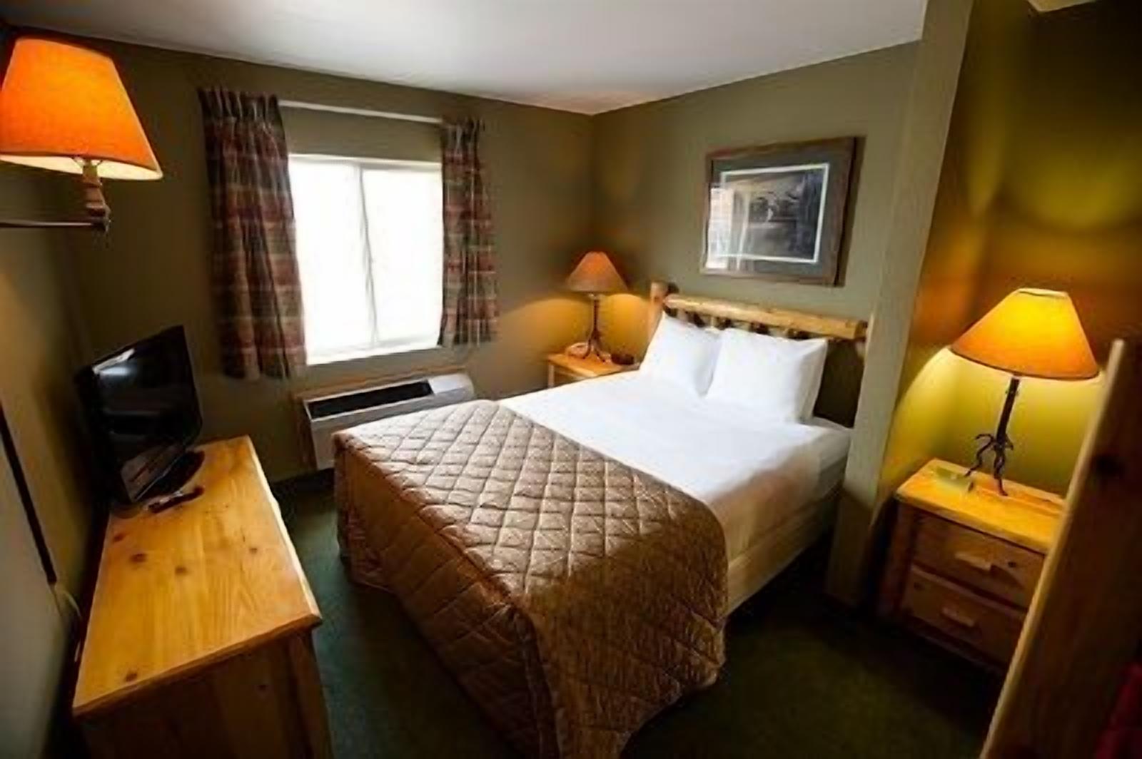 A hotel room with a bed, two bedside lamps, a television, a window with curtains, and a framed picture on the wall.