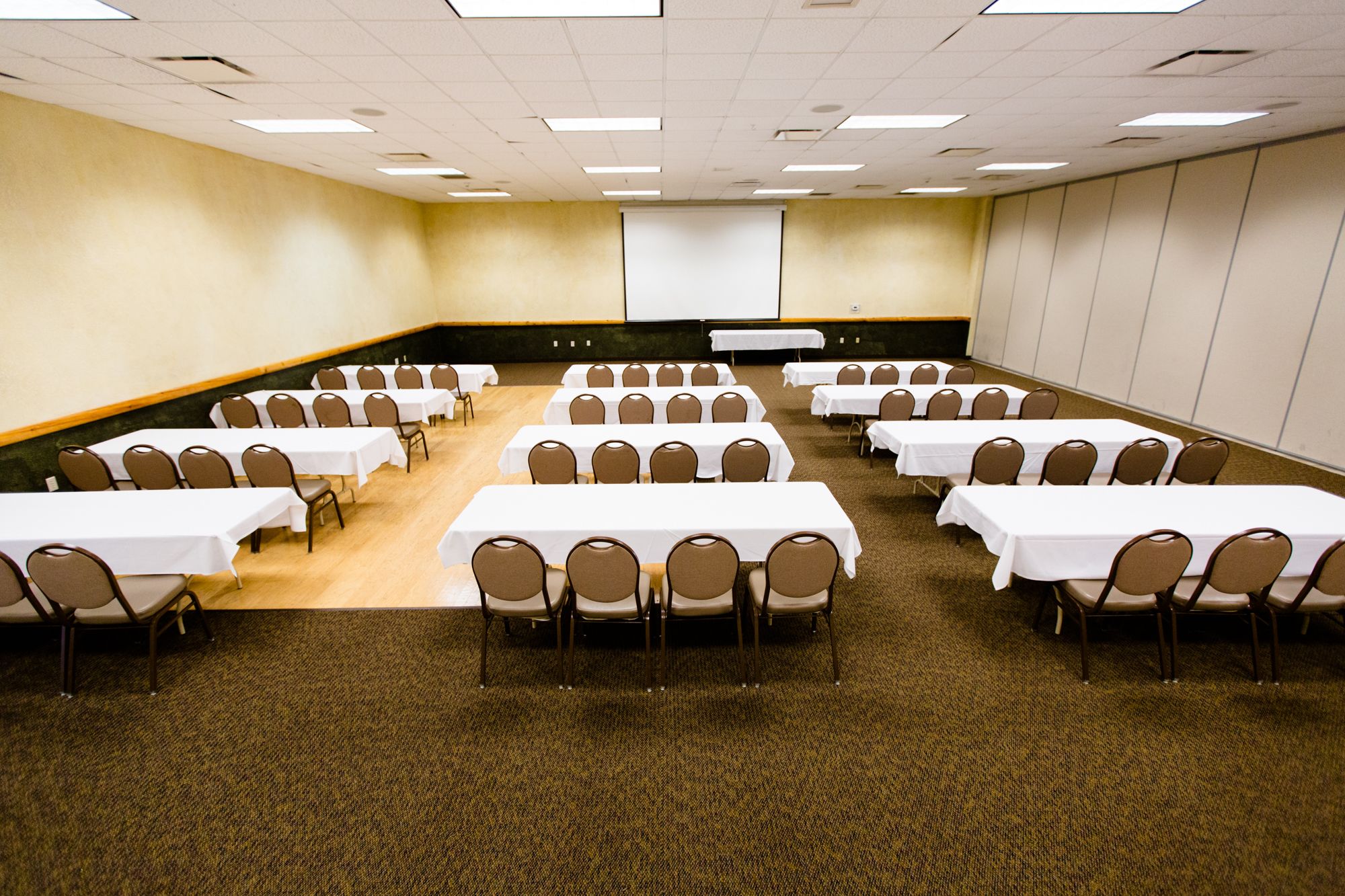 A conference room with multiple tables and chairs, a projector screen, and carpeted flooring.