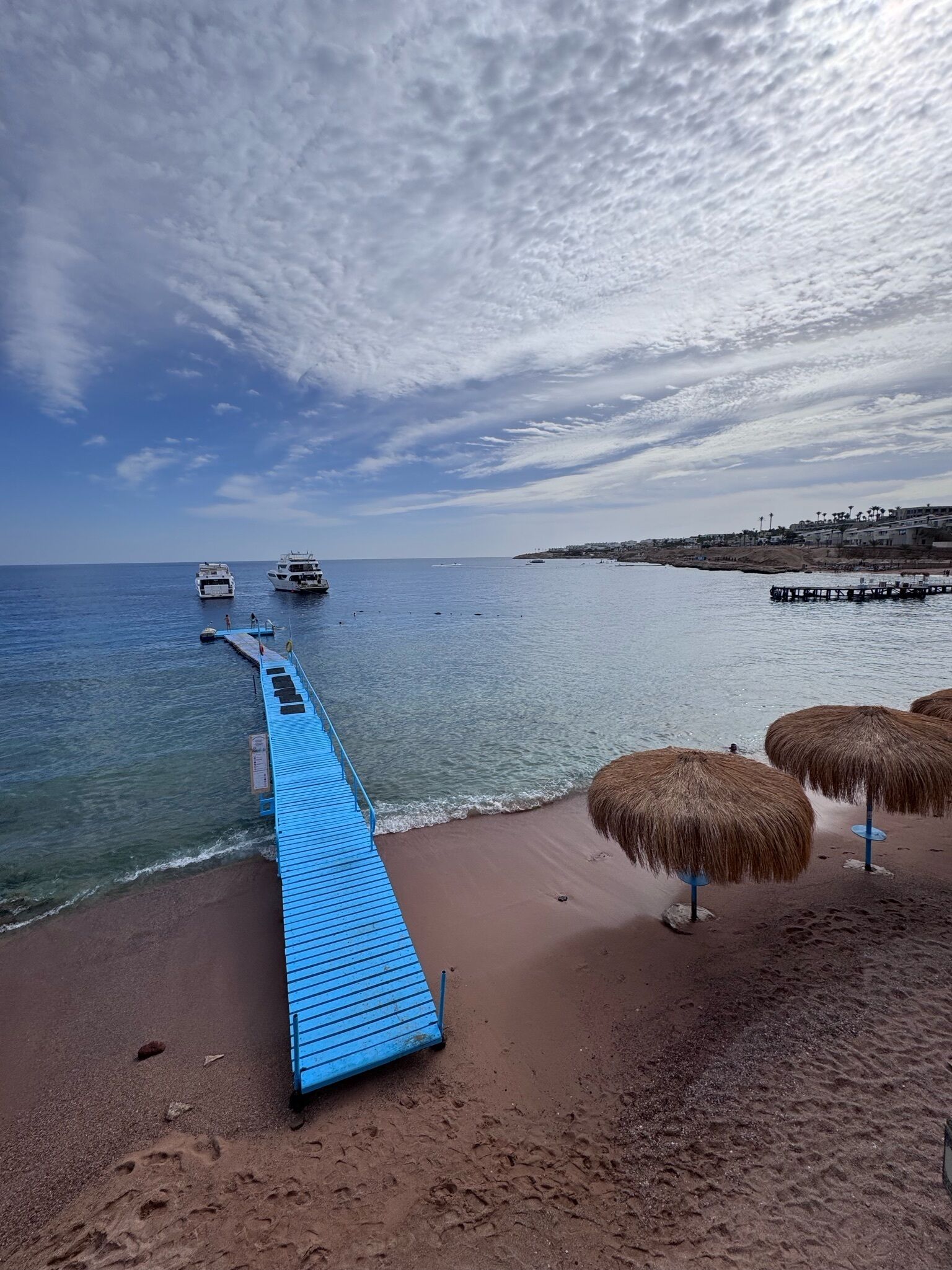 A beach with a blue jetty leading to the sea, thatched umbrellas, and boats in the distance.