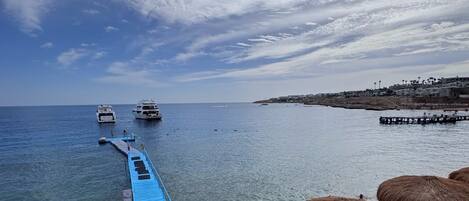 A beach with a blue jetty leading to the sea, thatched umbrellas, and boats in the distance.