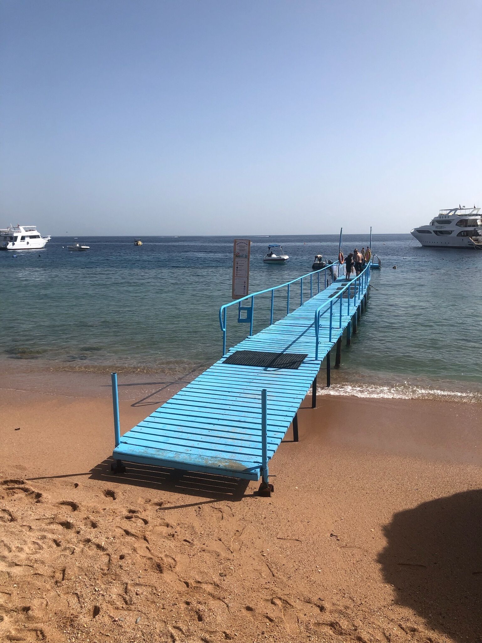 A beach with a blue pier extending into the water, boats, and a large yacht.