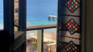 A view from a window overlooking a beach with a pier, a thatched-roof structure, and a person walking. - Umbi Sharks Bay Diving Village (Sharm El Sheikh)