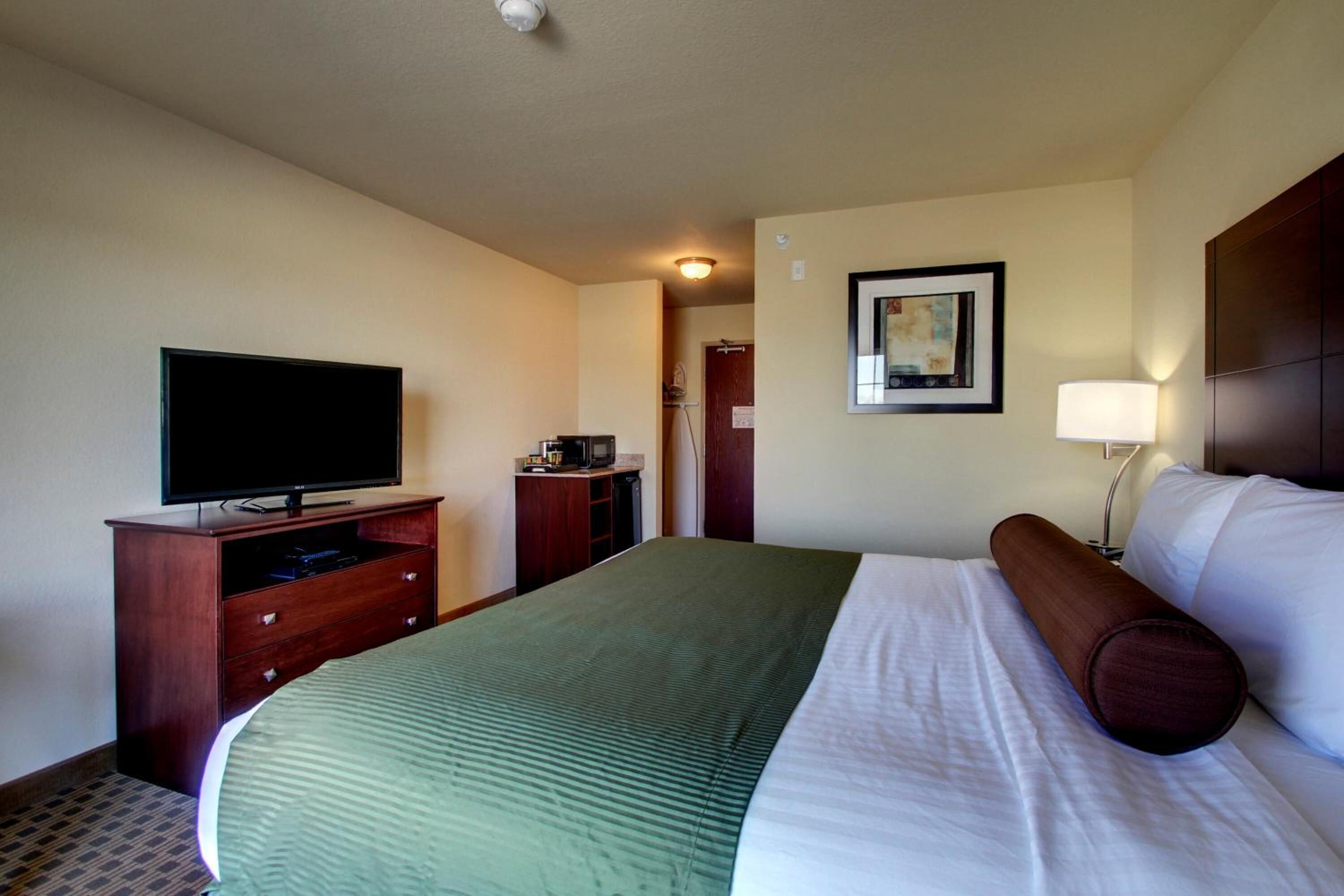 Een hotelkamer met een bed, een televisie op een houten standaard, een bureau met een lamp en een lijst aan de muur.