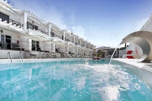 A swimming pool with lounge chairs and umbrellas, surrounded by a white building with balconies.