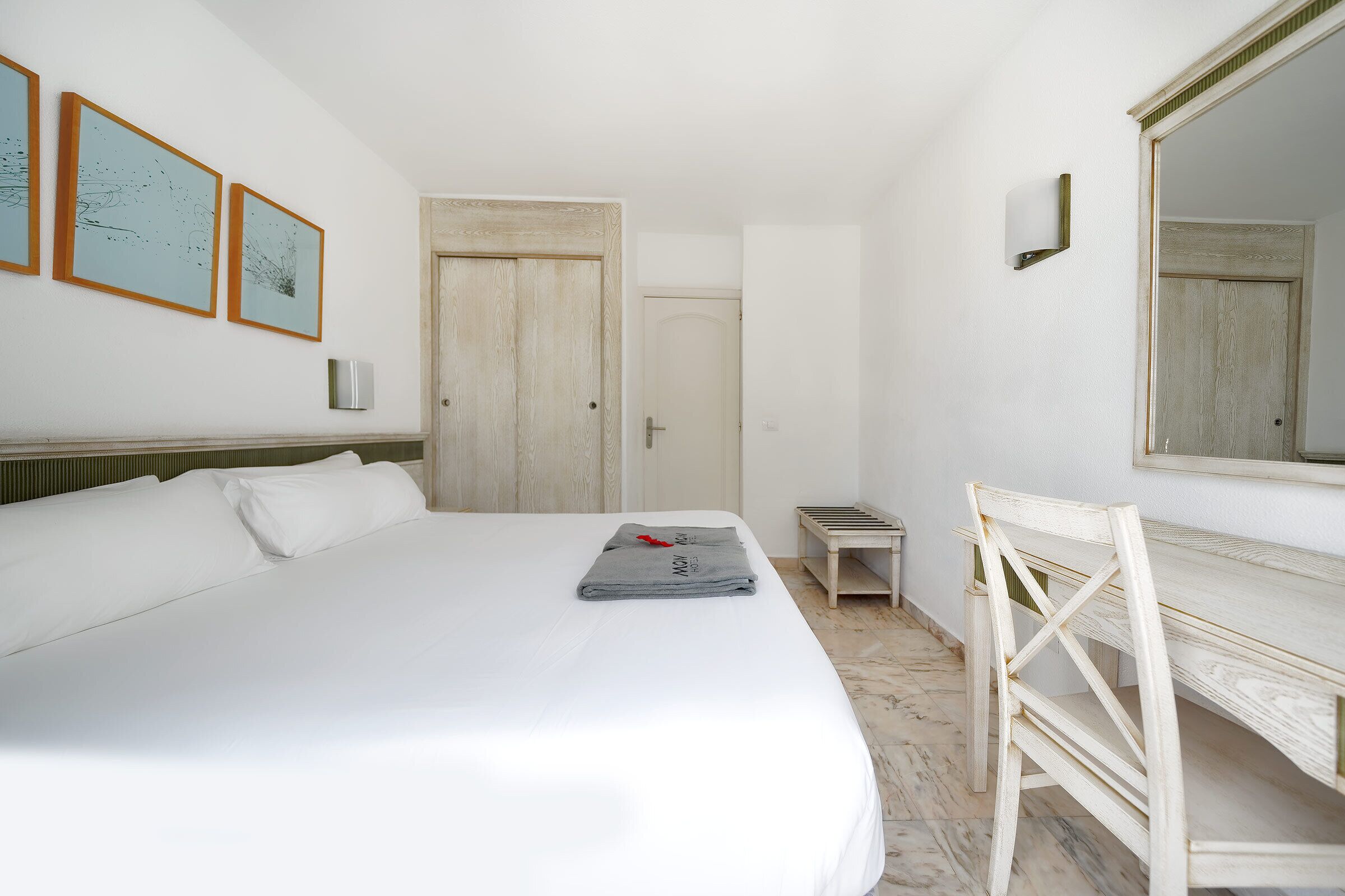 A hotel room with a large bed, a wooden desk, a small bench, and a mirror.