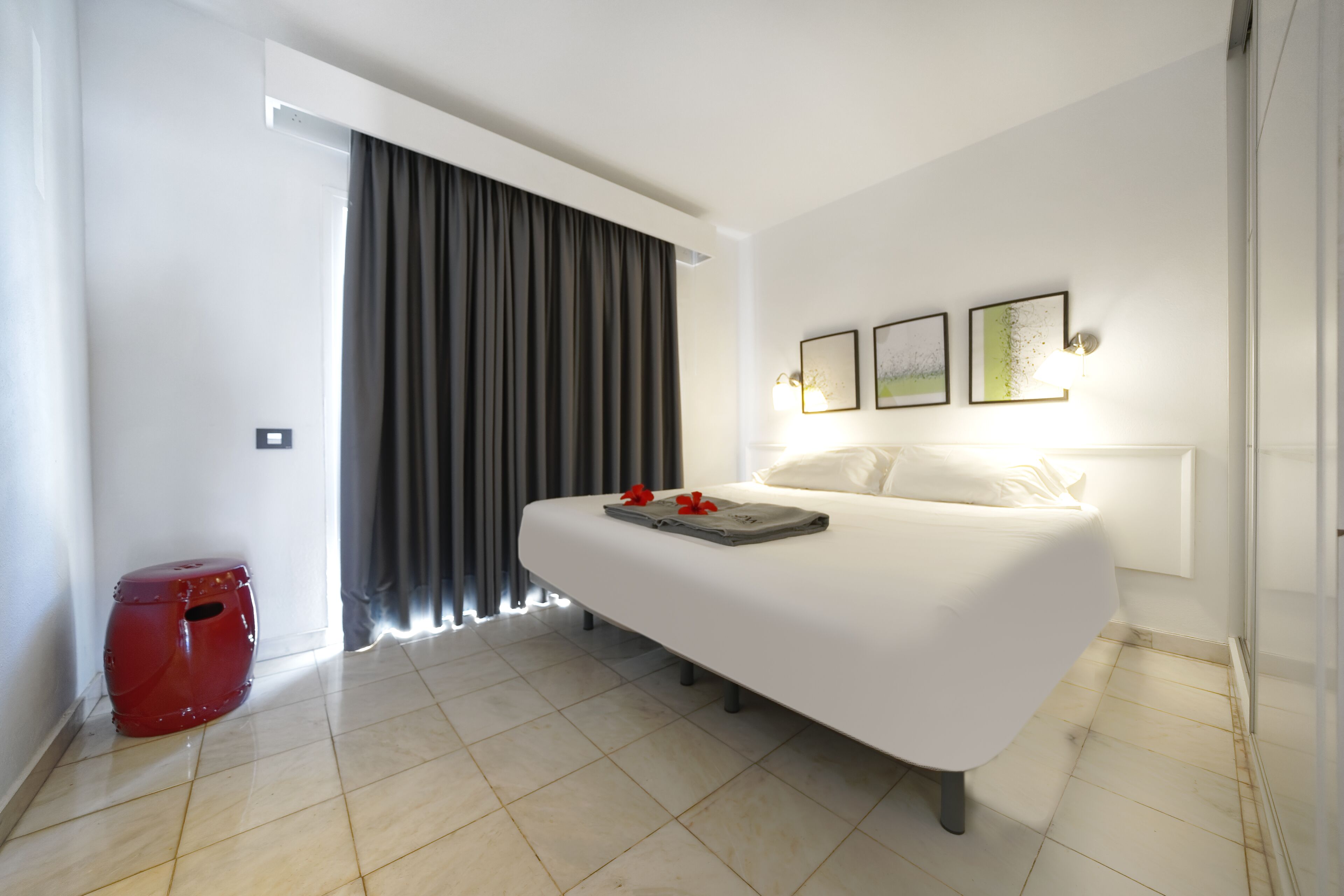 A modern hotel room with a large bed, a red stool, and a window with curtains.
