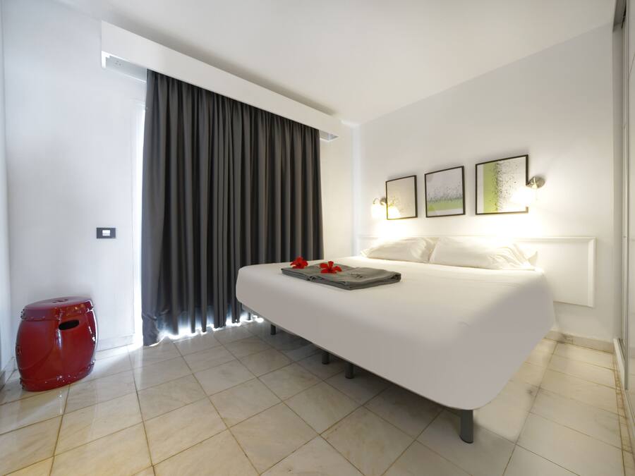 A modern hotel room with a large bed, a red stool, and a window with curtains.