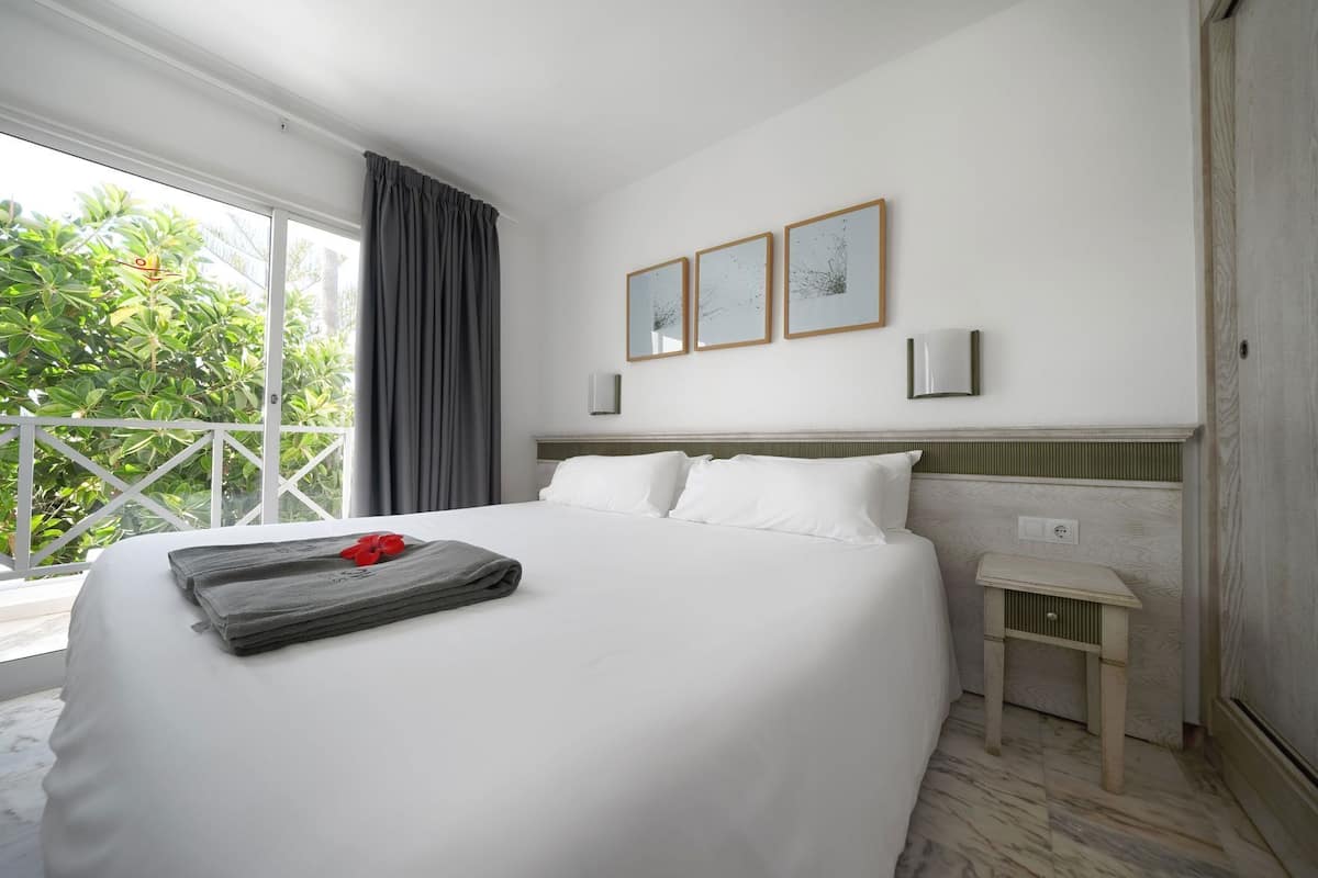 A hotel room with a large bed, a small bedside table, a window with curtains, and a balcony with greenery.