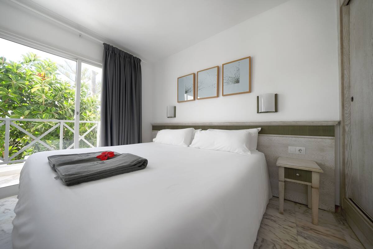 Une chambre d’hôtel avec un grand lit, une petite table de chevet, une fenêtre avec des rideaux et trois tableaux encadrés accrochés au mur.
