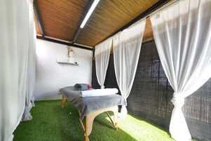 A room with a bed, white curtains, a small shelf with items, and a window with white curtains.