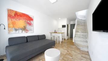 A modern living room with a grey sofa, a white coffee table, and a large abstract painting on the wall. A kitchen area with a wooden countertop and white stools is visible in the background.