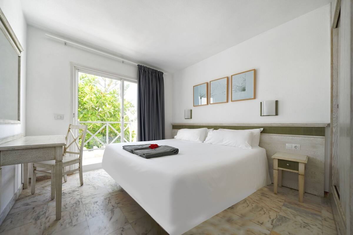Une chambre d’hôtel comprenant un lit, un bureau, une chaise, une petite table de chevet et un balcon avec vue sur la verdure.