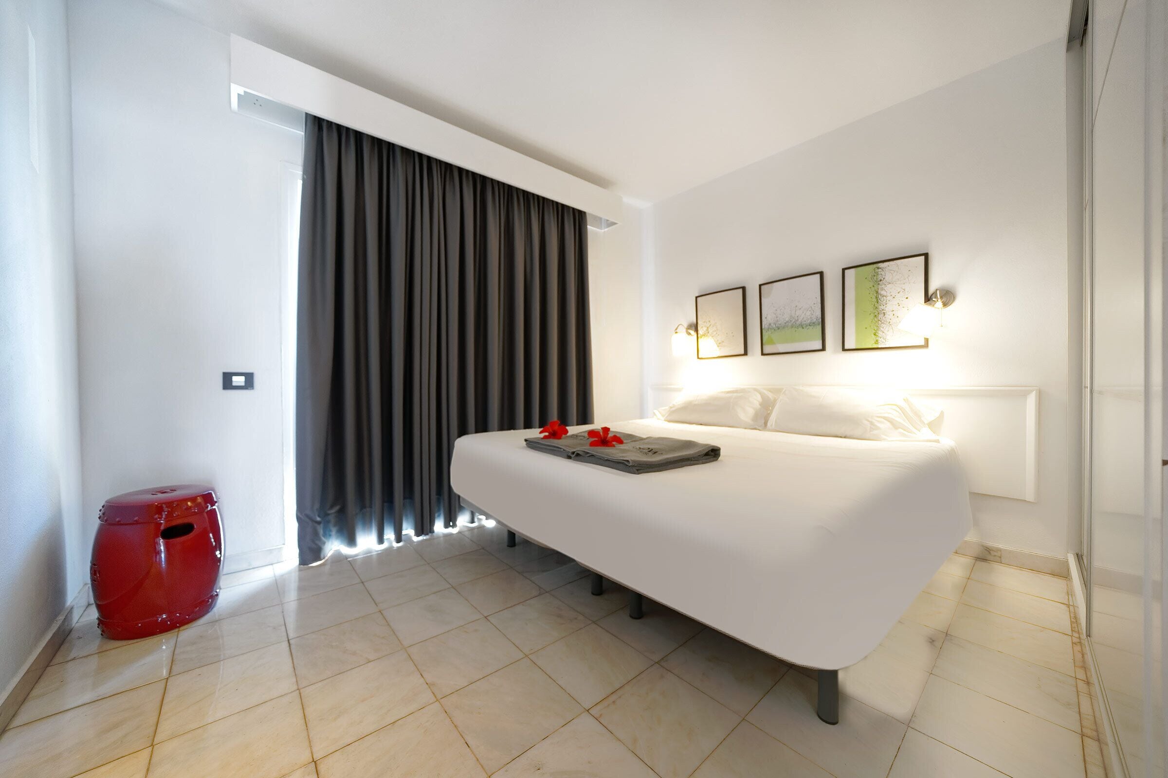 A hotel room with a bed, a red stool, and a window with curtains.
