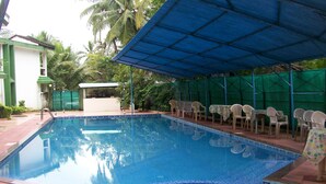 Una piscina con borde de azulejos, un área de estar cubierta con sillas de plástico y un edificio pequeño al fondo.