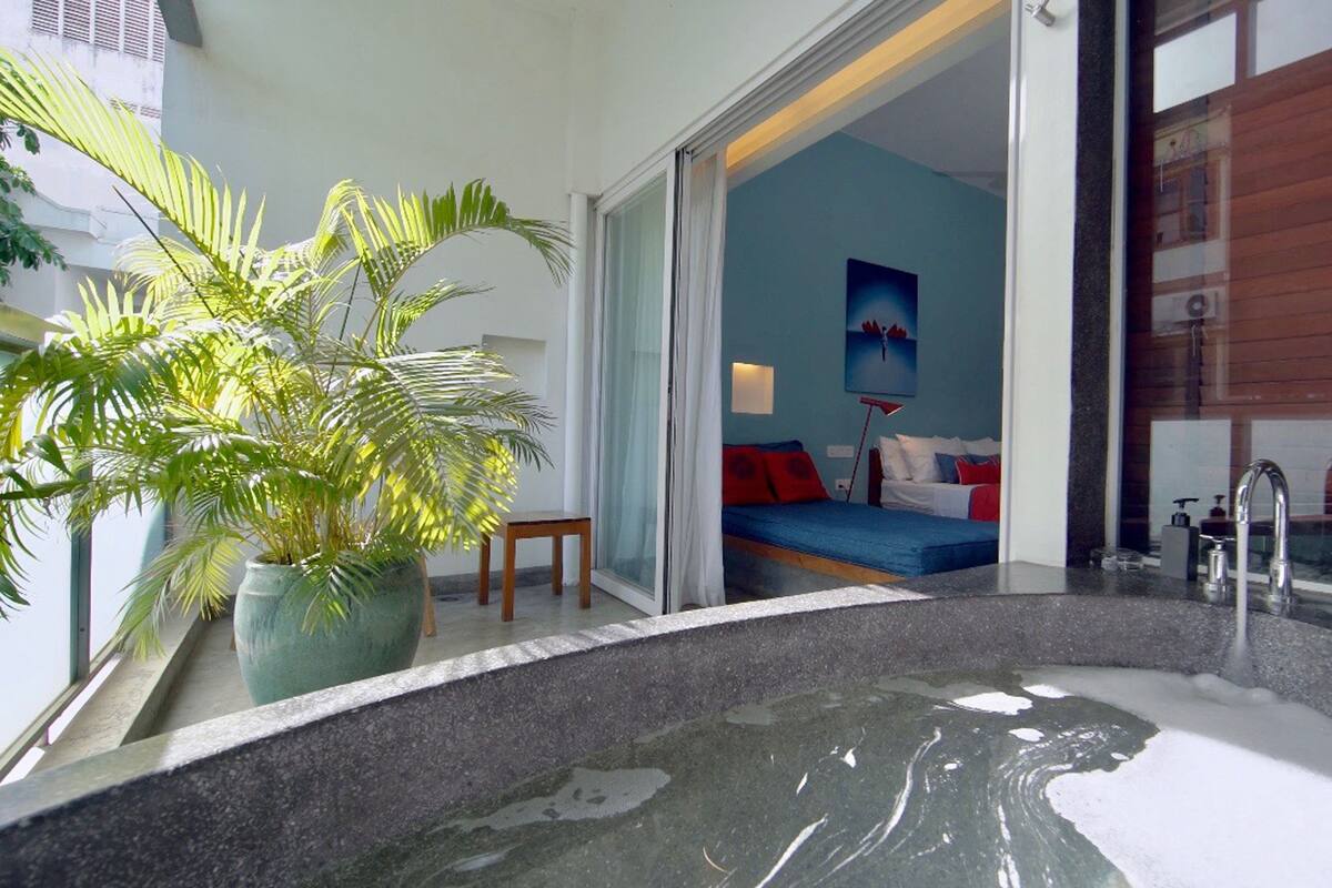 A modern bathroom with a large bathtub, a potted plant, and a view of a bedroom with a bed and a painting.