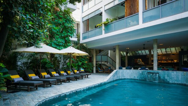 A pool area with lounge chairs and umbrellas, surrounded by greenery and a modern building structure. -  Sonitori Boutique Resort (Phnom Penh)