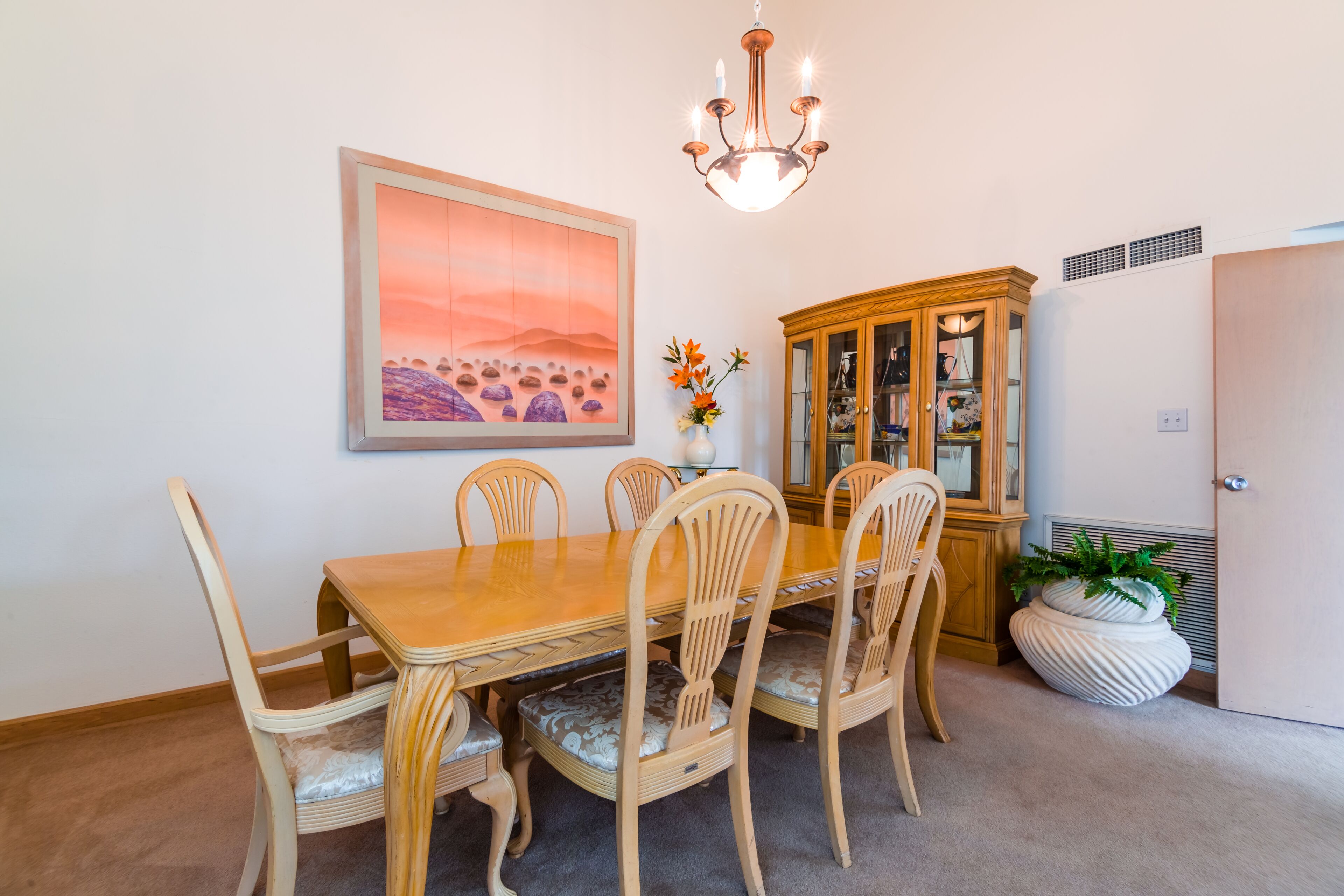 A dining room with a wooden table and chairs, a chandelier, a cabinet, and a painting on the wall.