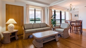 A living room with a sofa, two armchairs, a coffee table, a lamp, and a dining table with chairs. There are large windows with curtains, a plant, and a chandelier. - Alupang Beach Tower (Tamuning)
