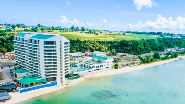 A high-rise hotel with multiple floors and a rooftop pool, situated by a beach with clear turquoise waters. - Alupang Beach Tower (Tamuning)