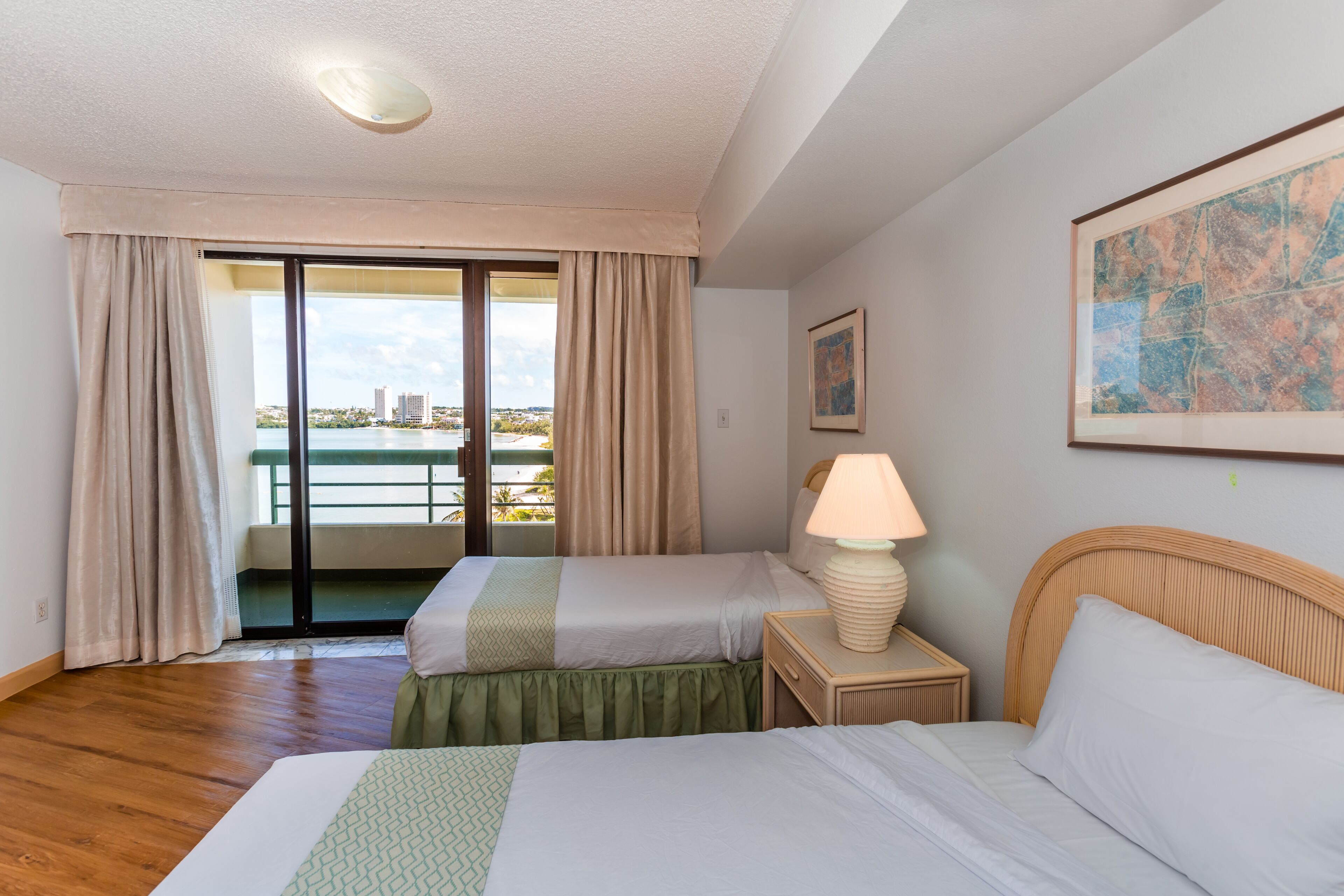 Une chambre d’hôtel avec deux lits, une vue sur la ville par une porte coulissante, une lampe sur une table de chevet et un tableau encadré au mur.