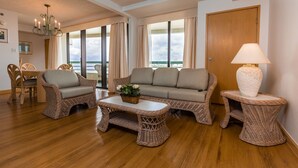 A spacious living room with wicker furniture, a chandelier, and large windows offering a view of the sea. - Alupang Beach Tower (Tamuning)