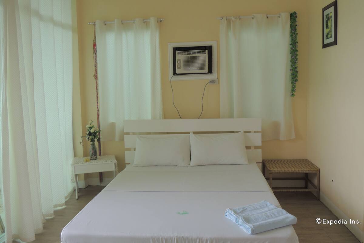 A hotel room with a bed, a bedside table, a small bedside table with a vase, a wall-mounted air conditioner, and a picture frame on the wall.