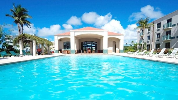 A swimming pool with a building in the background, palm trees, and lounge chairs. - Garden Villa Hotel (Tamuning)