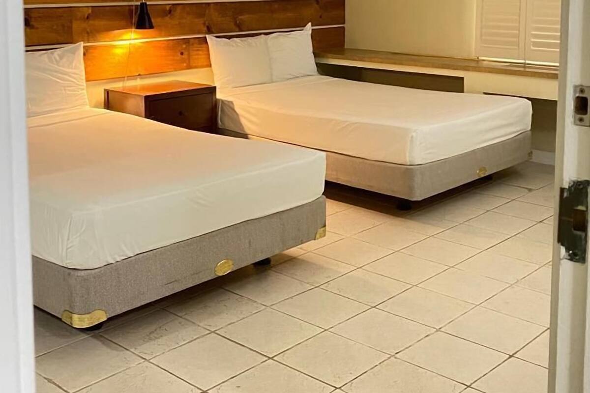 A hotel room with two beds, a wooden headboard, a bedside table with a lamp, and a window with blinds.