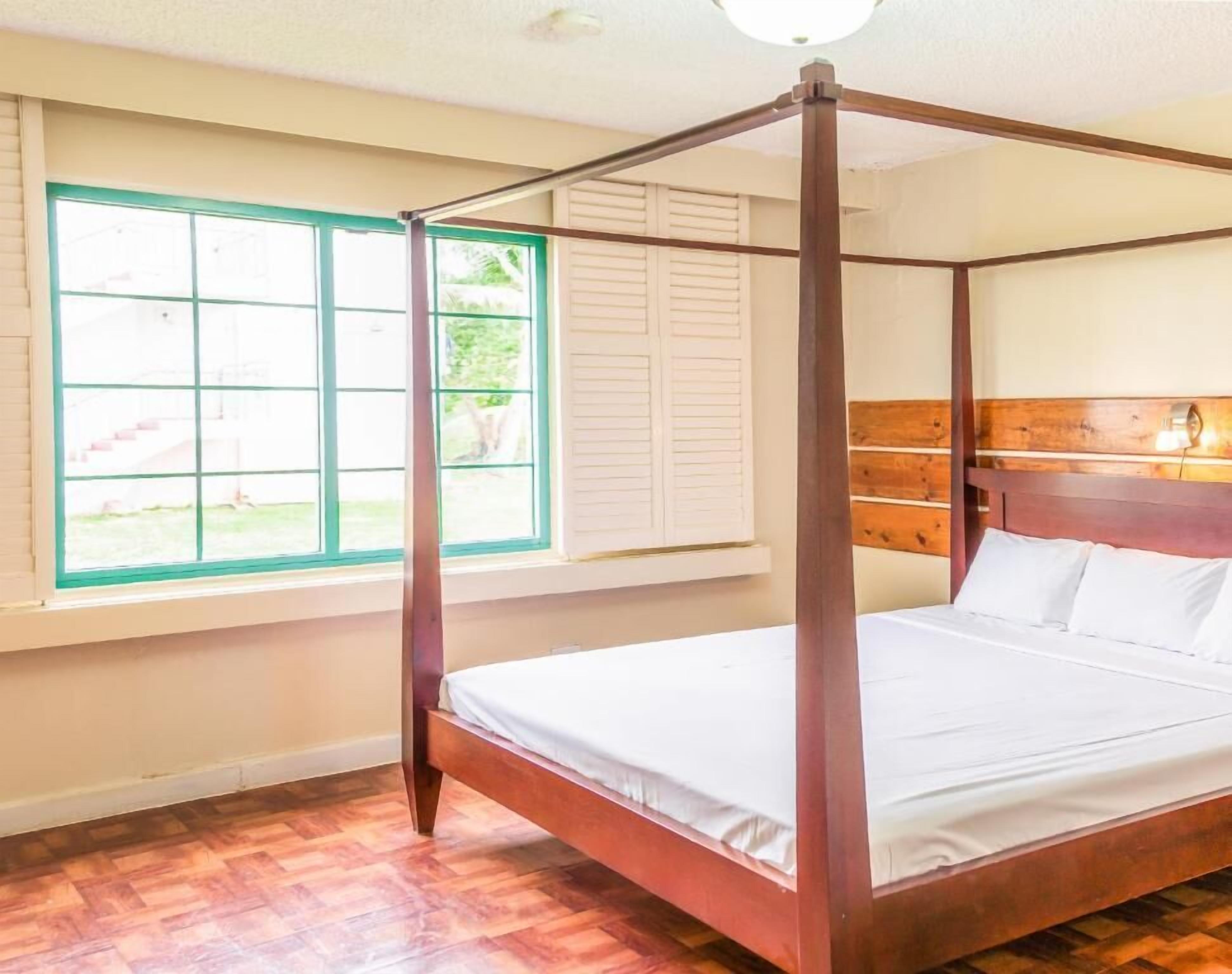 A four-poster bed with a wooden headboard in a room with wooden flooring and a window with white shutters.