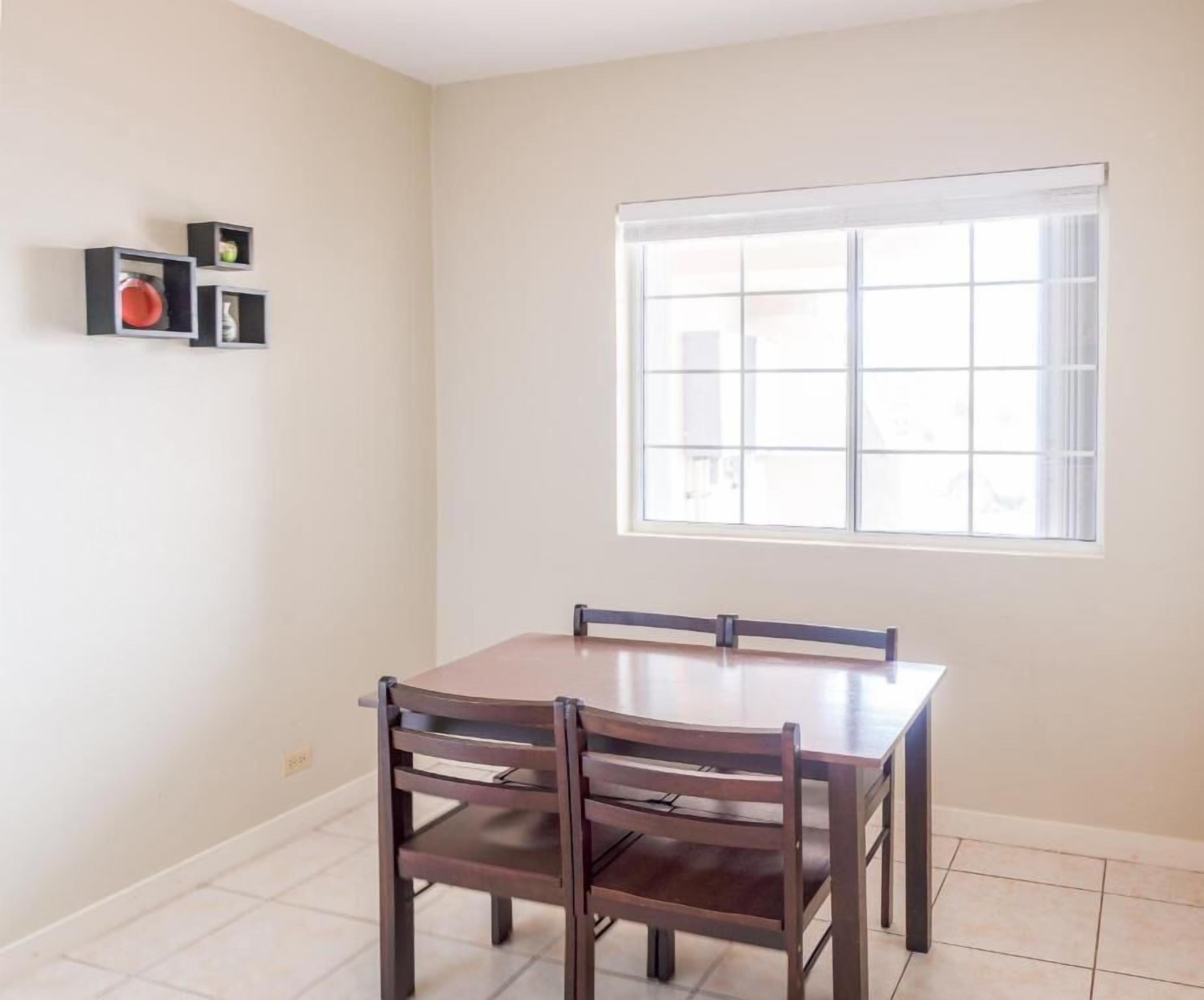 A dining area with a table and four chairs, a window with blinds, and a wall-mounted shelf with items.
