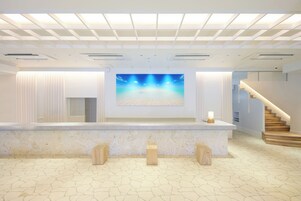 Zona de recepción con una gran pantalla digital mostrando una escena de playa, un mostrador de mármol y una escalera que conduce a pisos superiores.