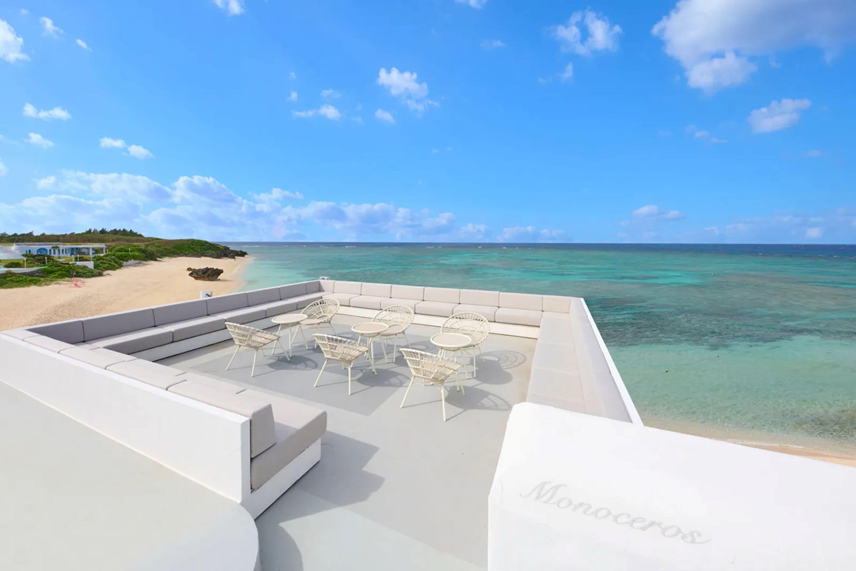 A rooftop terrace with a view of the ocean, white outdoor furniture, and a building in the background.