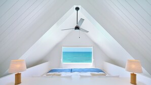 A bedroom with a sloped ceiling, a ceiling fan, a window with a view of the sea, and two bedside lamps. - Pricia Resort Yoron (Yoron)